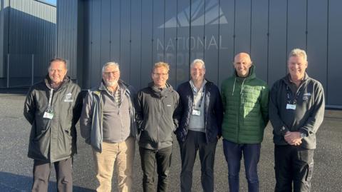 Six men standing in a line in front of a large door saying National Drone Hub.