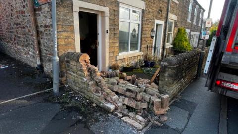 A front garden wall which has been knocked down - there are bricks all over the front yard and to the right there is a lorry parked on the pavement. 
