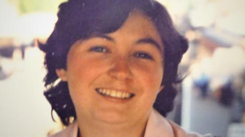 A woman with mid length brown hair smiles towards the camera, the collar of the pink top or coat she is wearing is barely visible. The background is out of focus