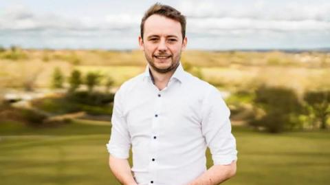 Chris Curtis, the Labour MP for Milton Keynes North. He is smiling to camera. He has short light brown hair, swept to one side. He is wearing a fitted white shirt and is stood outside with hills behind him in the background. 