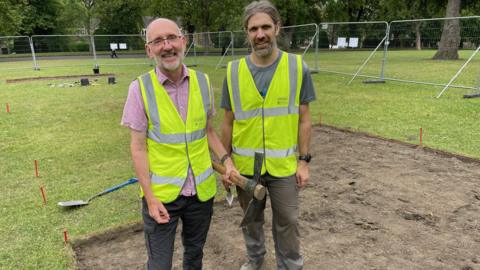 Two men wearing high visibility yellow jackets stand in a dug up part of a field which is fenced off. They both carry digging tools and are surrounded by others on the ground.