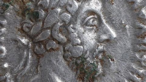 A close-up of a silver denarii showing a profile of the emperor Marcus Aurelius. He  is wearing laurel leaves, and has a beard. The coin shows some signs of green around his beard. 