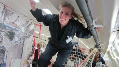 Susan Charlesworth has long blonde hair and is wearing a green jumpsuit. She is in an anti-gravity plane and floating near the ceiling.
