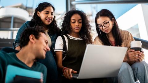 Four students, three women and one man, looking at a laptop. One is holding a coffee cup