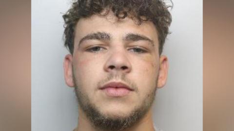 A custody photo of a man with short dark curly hair, a short beard and dark eyes. The background behind him is grey.