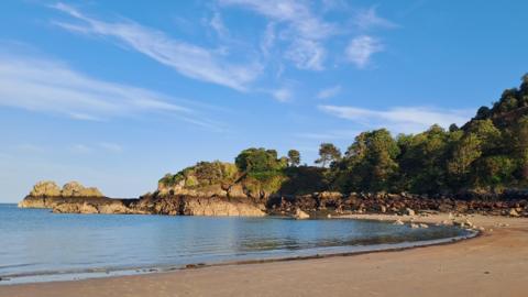 Gorey in Jersey. A sandy beach with blue clear water. The sky is blue with white clouds. In the background are trees.