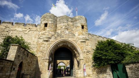 Imposing, high stone castle walls and a large gatehouse with an arched entrance and two circular towers above it topped with flagpoles flying the flags of Lincolnshire and the Union. People can be seen walking under the archway. The sky is blue.