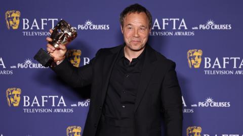 Stephen Graham wears a black suit and shit and is smiling holding a gold BAFTA award. He is stood in front of a purple backdrop which is covered in the BAFTA logo. He has short dark hair