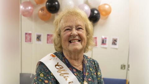 A head and shoulders shot of Janet Harley smiling at the camera. She has short, blonde hair and is wearing a green, floral dress and a pink and gold sash. Black, orange, pink and white balloons are stuck to the cream-coloured wall behind her. 