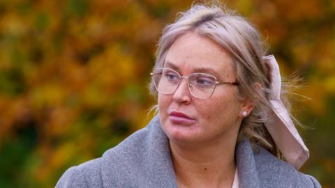 A blonde haired woman, with her hair tied back in a light pink bow. She wears gold rimmed glasses 