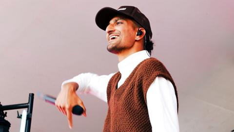 Deeps performing at Radio 1's Big Weekend, wearing black hat, a white long sleeved top with a brown knitted vest jumper over it. He is holding a microphone in his right hand.