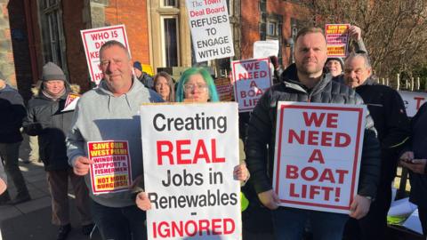 Men and women stand with placards that read "creating real jobs in renewables - ignored", "we need a boat lift" and "save our West Pier, windfarm support and fishermen, not more burgers".