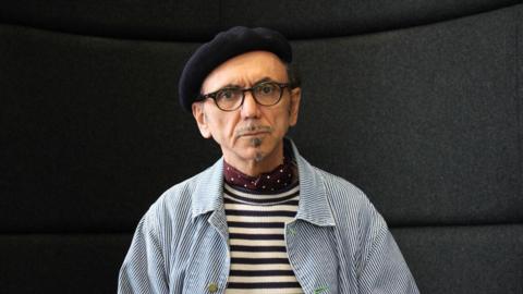 A man with dark glasses, a small goatee beard a black beret and serious expression in a blue and white striped jacket and black and white striped tshirt in front of a black background