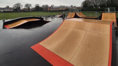 A skate park with different levels of beige ramps with a red exterior on black flooring. In the distance is a field with houses around it.