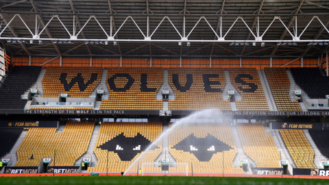 A generic view of Molineux stadium. Rows of seats in yellow and black form the word "Wolves" alongside two wolf logos. A sprinkler in the foreground is watering the turf