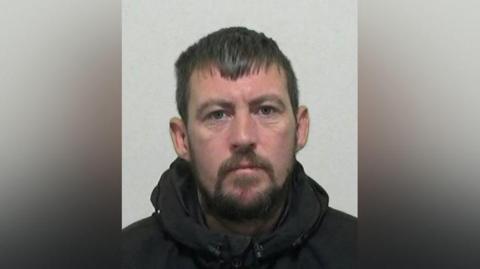 Police mugshot of Liam Doeg. He has black hair and a black beard and is wearing a black hooded coat.