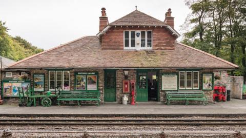 Woody Bay Station viewed from across the railway tracks. It is in the style of a Swiss chalet and has two chimneys, a slanted roof and green doors. There are green benches outside it and old-style luggage carrying trolleys. There is also a post box built in to the front wall. There are trees beyond.