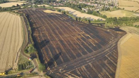 A large blackened field, with trees all around the perimeter, and homes to the top right, and a pylon to the top left. Other fields, that are not burnt are around the area. 