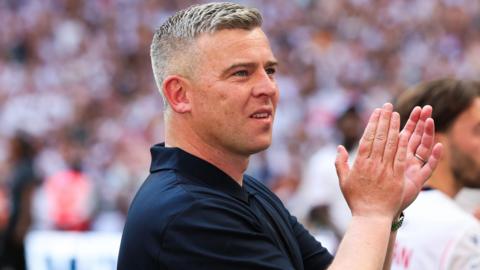 Steven Schumacher, wearing a navy blue polo shirt, claps his hands in celebration after Bolton beat Stockport in the League One play-off final in May 2026