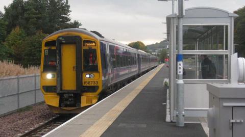 A train sits at the platform while one person waits in a shelter on the Borders Railway