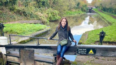 A woman sits on a wooden canal lock with the waterway visible behind her. She has long dark hair, and is wearing a grey jacket, blue jeams and brown leather boots.