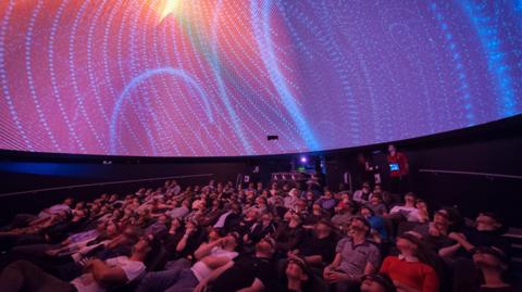 An audience looking up at the ceiling in a planetarium. The display is pink and there are blue dots all over it.