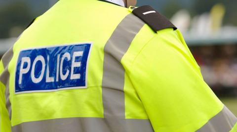 The back of a police officer who is wearing a bright yellow high-vis coat with the word "police" in white capital letters on a blue rectangular panel on it