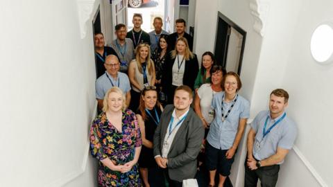 A crowd of smiling staff and councillors looking up the stairs of a new homeless hub. The door is open behind them.