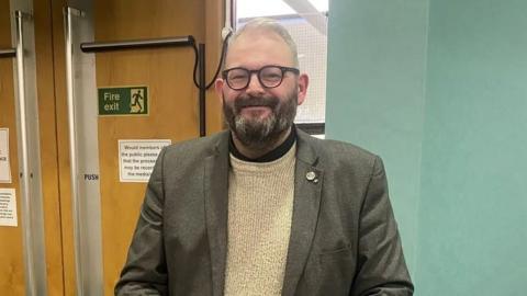 Mike Young has a dark beard that is greying and grey hair. He wears glasses and smiles at the camera. He is wearing a brown suit jacket and beige jumper.
