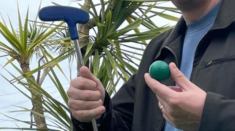 A man holding a green ball and a rubber-headed club at a crazy/adventure golf course. There is a palm tree in the background.