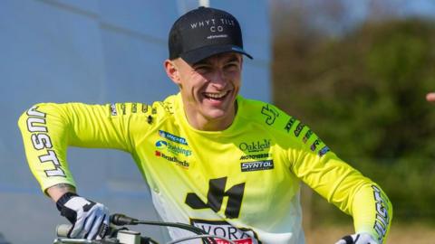 Calum, a man, wearing a black cap and a light green long-sleeve shirt. He is sitting on a motorbike and smiling but looking away from the camera.