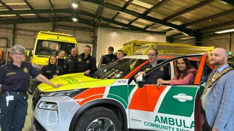A group of ambulance staff in green uniform stand by a 4x4 vehicle with orange and green panelling and a yellow bonnet. A man in a light blue suit stands to the side of the vehicle wearing a large gold chain. A woman in a pink suit leans through the vehicle's window to smile at the camera. 