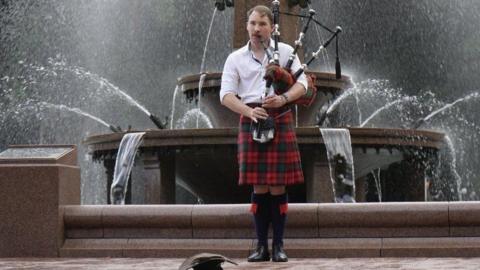 A man with a red kilt and white shirt playing the bagpipes in front of a fountain