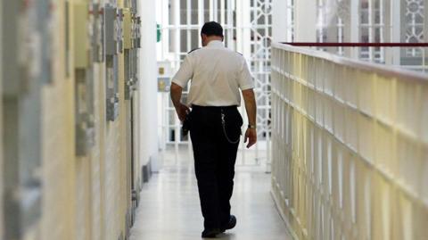 A prison officer walks down the corridor of a prison with his back to the camera. 