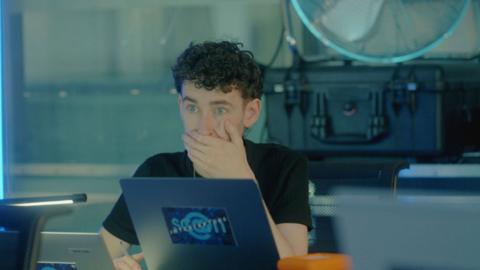 Man sat in front of a laptop holds his hand against his mouth. He looks shocked.