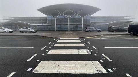 A zebra crossing leading up to a small glass airport building on a foggy day.