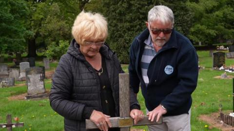Liz and Malcolm Hare are standing at Pete Wilson's grave with their hands on the wooden cross. They are both looking down at the grave.