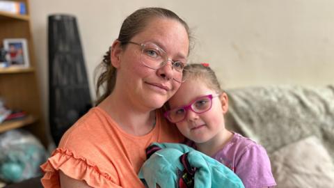 Kelly has tied back dark hair. She is wearing glasses and has nose piercings. She is wearing a coral coloured t-shirt with ruffled sleeves. Her daughter Connie is sitting on her lap and their faces are close together. Connie has her hair tied back and is wearing pink framed glasses. She is holding a blue soft toy.