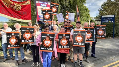 A group of about 20 people standing on a roadside holding placards saying "NHS Pay for NHS Jobs". Some of them wear NHS uniforms. Behind them is a sign for Airedale General Hospital.