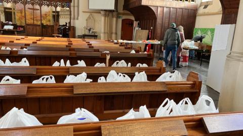 Dozens of food parcels in carrier bags line wooden church pews.