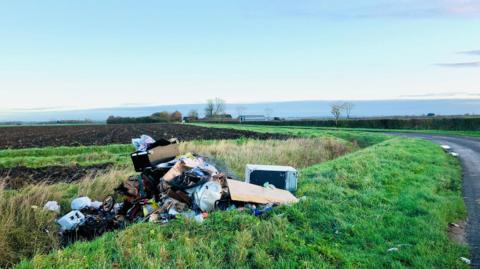 Pile of fly-tipped waste on a grass verge beside a road.