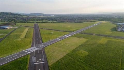 A small airfield shown from above, with two runways crossing. Countryside can be seen in the distance, and there are no planes on the tarmac. 