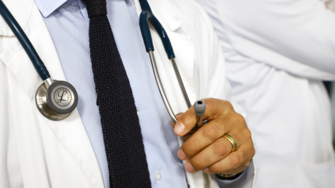 A generic picture of a doctor wearing a shirt and tie and a white jacket. He has a stethoscope around his neck, which he is holding. His face cannot be seen.