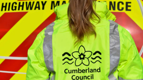 A female Cumberland Council wearing a yellow hi-vis jacket with the words Cumberland Council and its flower logo stands with her back to the camera, with the yellow and red lines of a council Highway Maintenance vehicle in the background