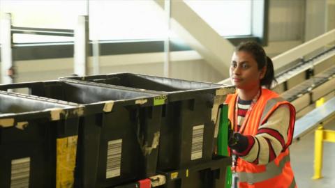 A woman in a orange hi-vis vest pushing a row of black boxes through a warehouse.