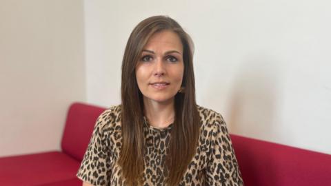 Lucy Domaille - A woman with long brown hair, brown eyes and a leopard print top on looking at the camera. She is sitting on a red sofa. 