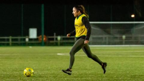 A women stands on a football pitch kicking a football. It is very dark and she is wearing a yellow T-shirt and black leggings