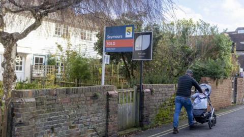 A white terrace behind a low wall. Two real estate agent signs are in place beside a wooden gate in the wall, while a man pushes a pram up the hill.