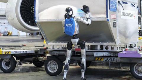 A blue, silver and black robot pushes a cargo truck at an airport, with a plane engine visible in the background