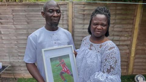 Peter Oppong and Mintaa Okyere wearing white in front of a garden fence and holding a framed photograph of their son Othneil playing football
